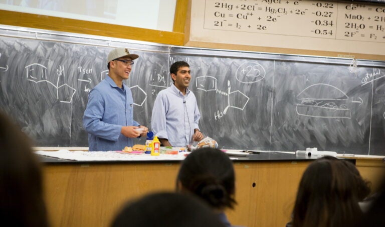 Two male instructors stand in front of a class with hamburger ingredients on a desk in front of them and a diagram of a molecule and a hamburger on a blackboard behind them.