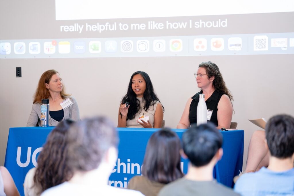  Biotechnology and Society Cluster teaching team members Michelle Rensel, Kyla Yein, and Chloe Bell-Wilson seated at a raised table in a lecture hall.