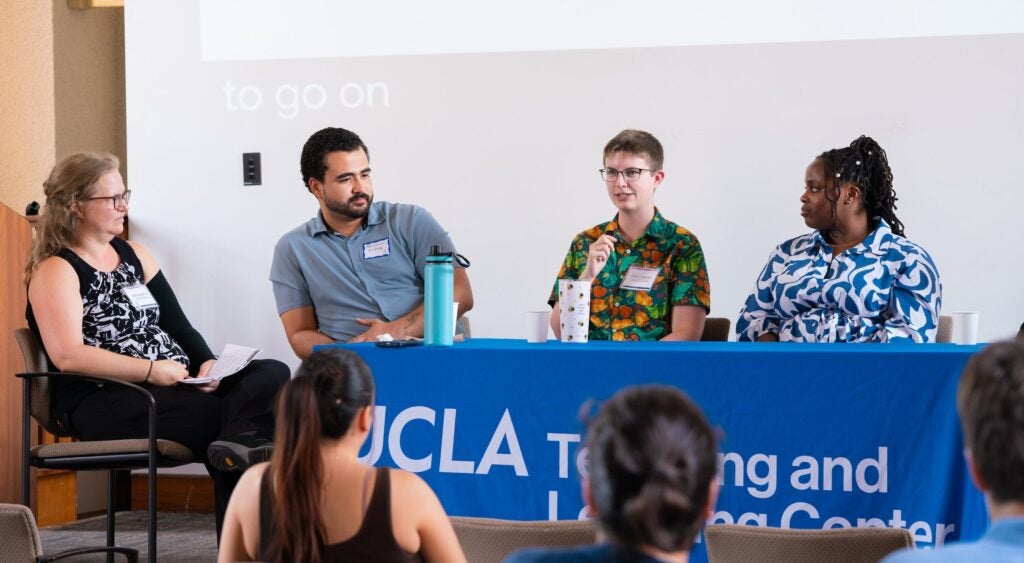 Four graduate students and postdoctoral scholars in conversation at the TA and Psotdoc Teaching Conference at a raised table with a blue tablecloth in a lecture hall.