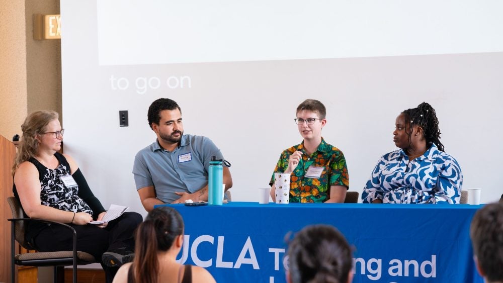 Four graduate students and postdoctoral scholars in conversation at the TA and Psotdoc Teaching Conference at a raised table with a blue tablecloth in a lecture hall.