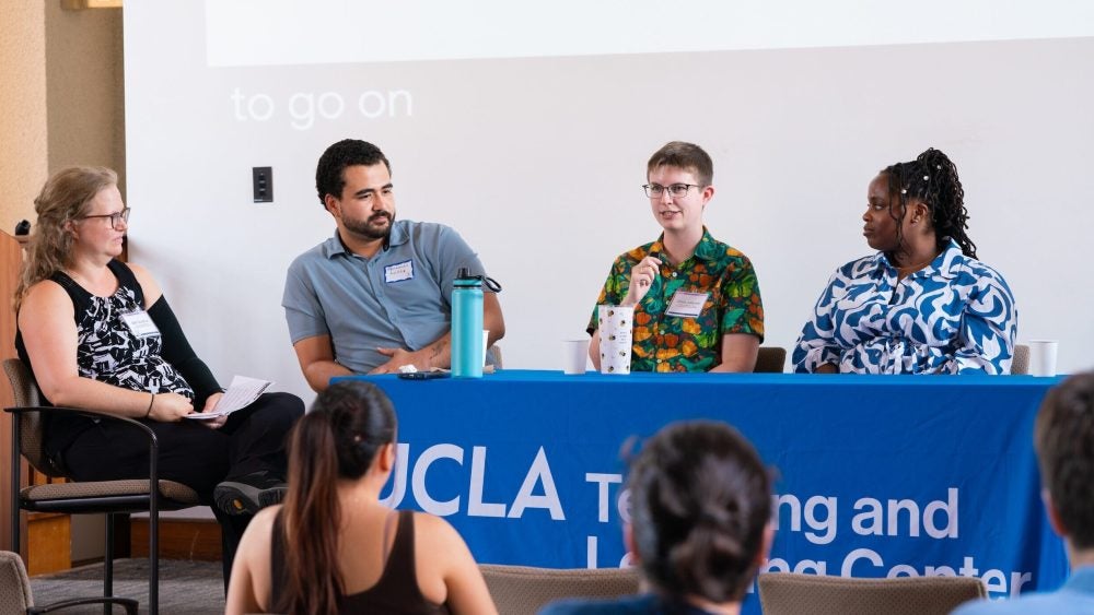 Four graduate students and postdoctoral scholars in conversation at the TA and Psotdoc Teaching Conference at a raised table with a blue tablecloth in a lecture hall.