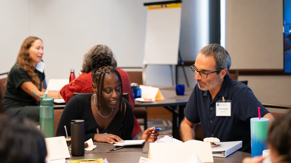 Two participants sit at a table and speak to each other while reviewing a notebook