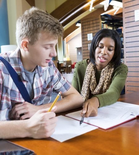 Teen or young adult African American college girl is tutoring a teenage Caucasian high school boy while they sit at table in modern school or public library. Tutor is explaining class assignment while student looks at paper. They are wearing trendy casual cothing.