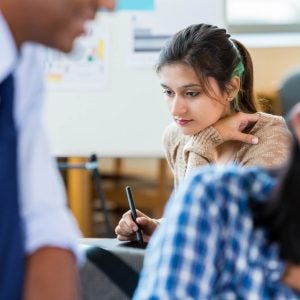 Pretty young Hispanic woman daydreams as she works on project in creative design firm. African American manager works with young Asian employee in the foreground.