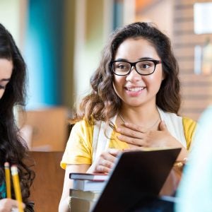 Trendy teen or young adult Hispanic woman is smiling and talking to diverse group of girl friends while studying for exam in college or public library. Students are using laptop computers and taking notes.