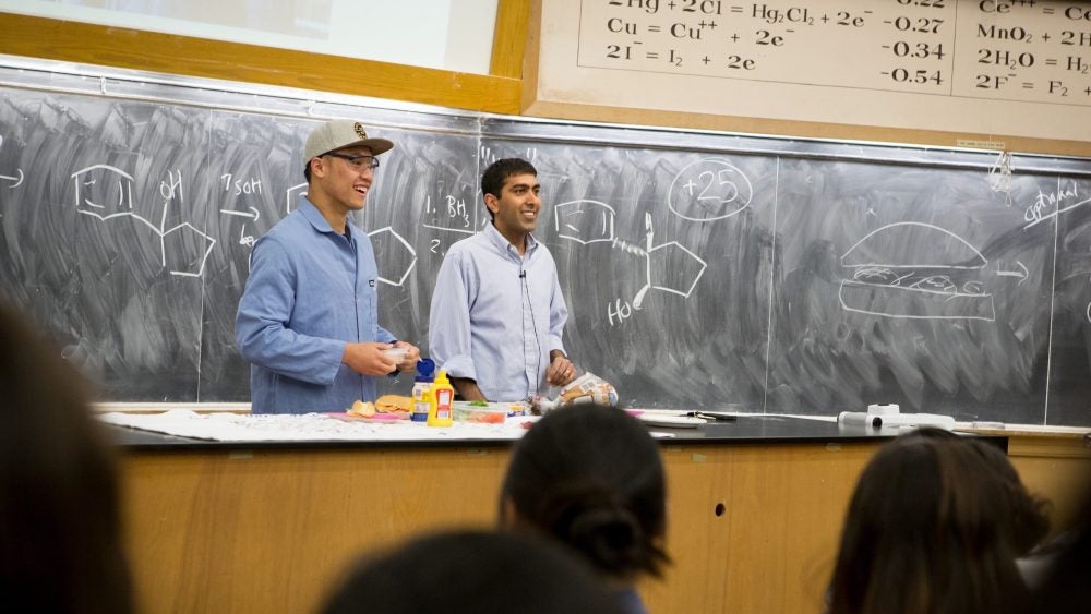 Two male instructors stand in front of a class with hamburger ingredients on a desk in front of them and a diagram of a molecule and a hamburger on a blackboard behind them.